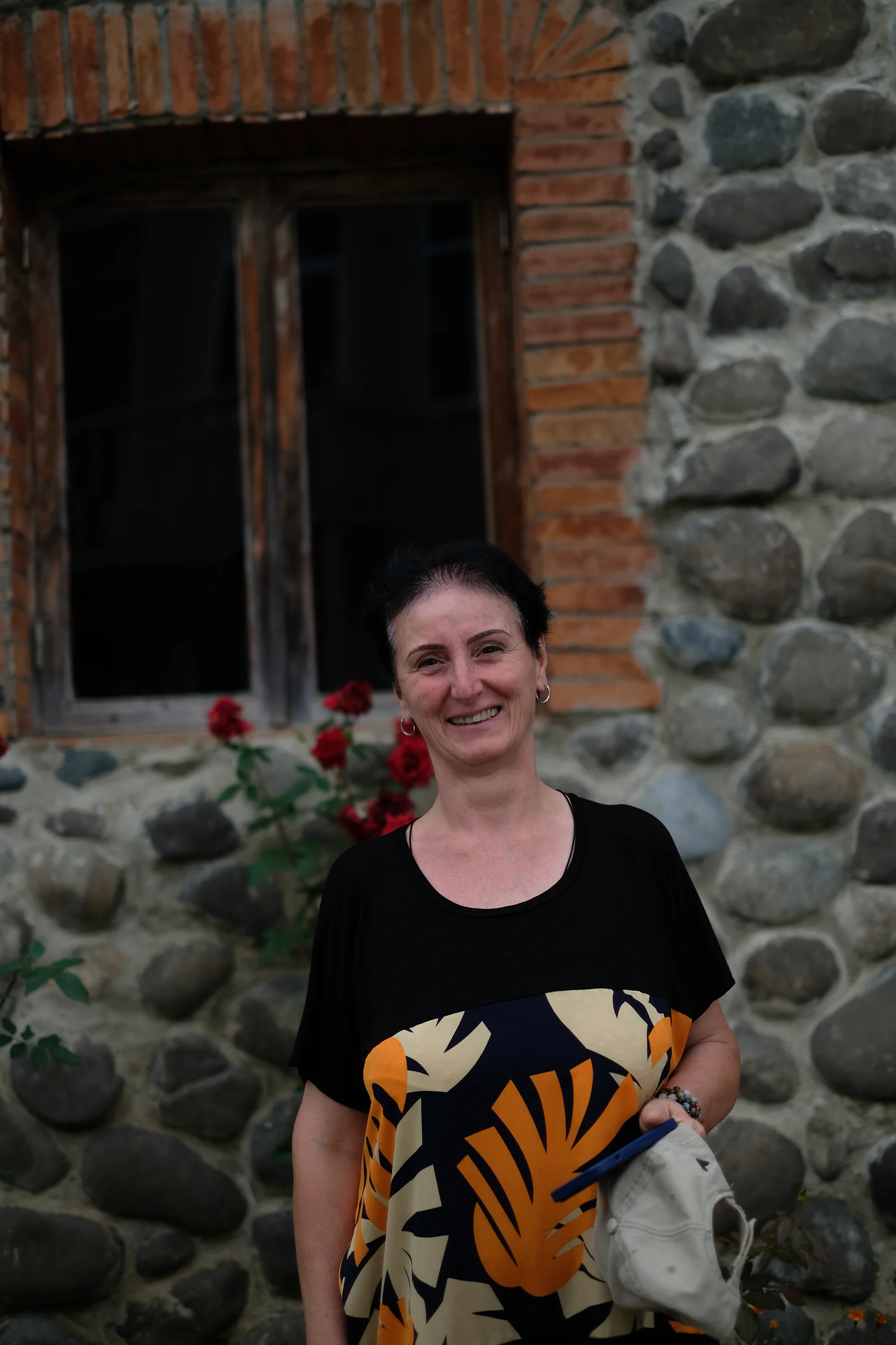 Baia's mother smiling outside the winery, in front of a stone wall and red roses.