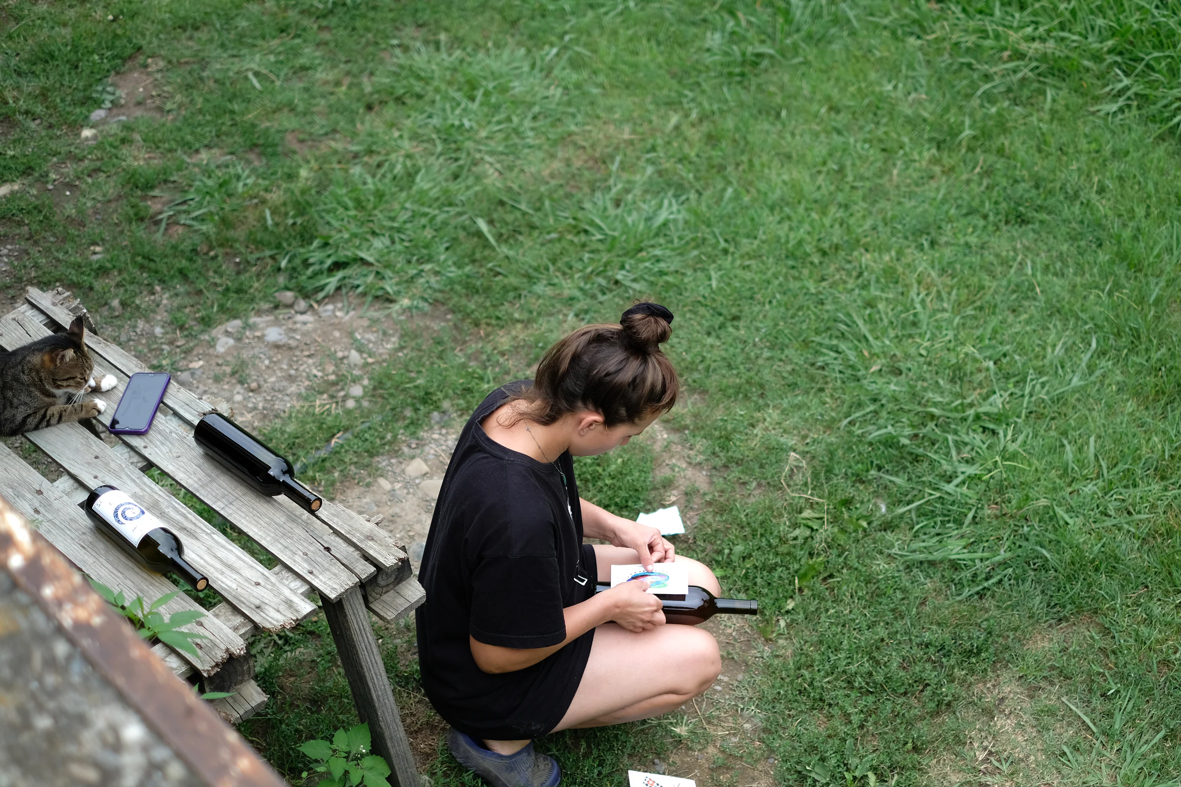 Ének Peterson kneeling beside a wooden bench as she attaches a label to a wine bottle, with a cat and bottles nearby.