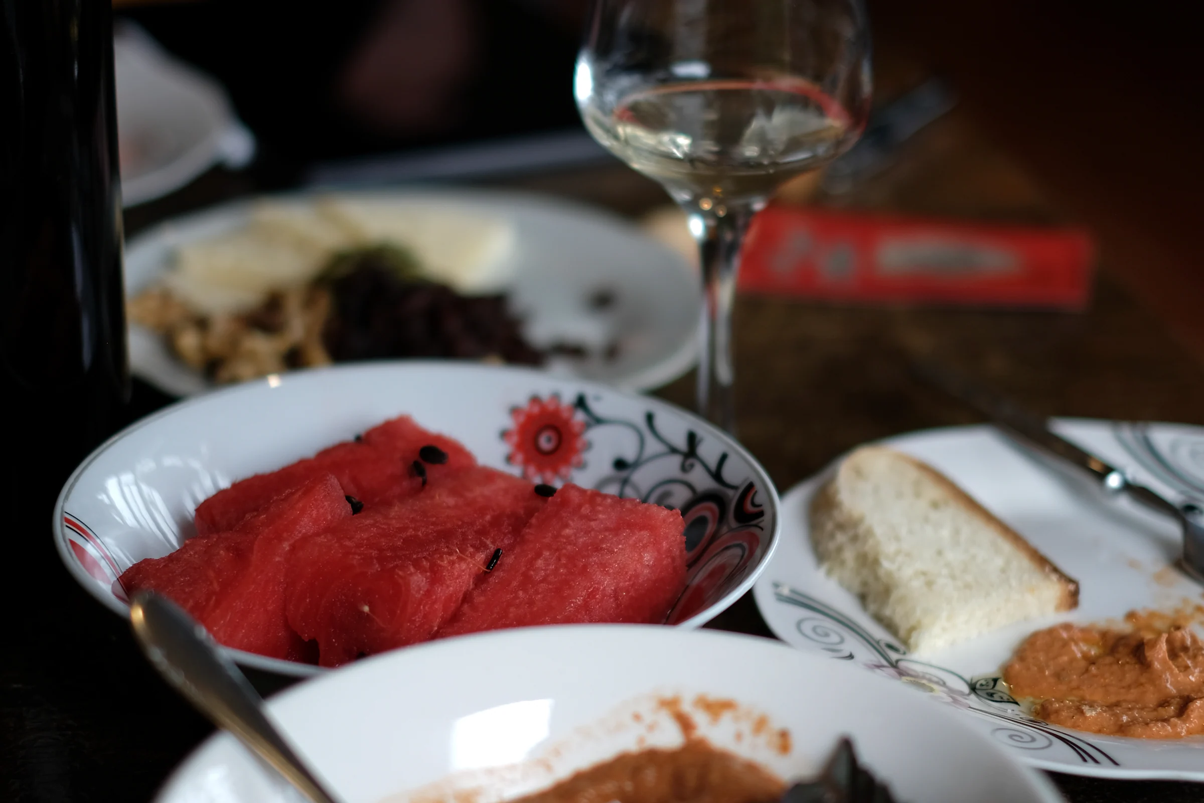 A bowl of sliced watermelon on the table beside a wine glass, bread and the red sauce.