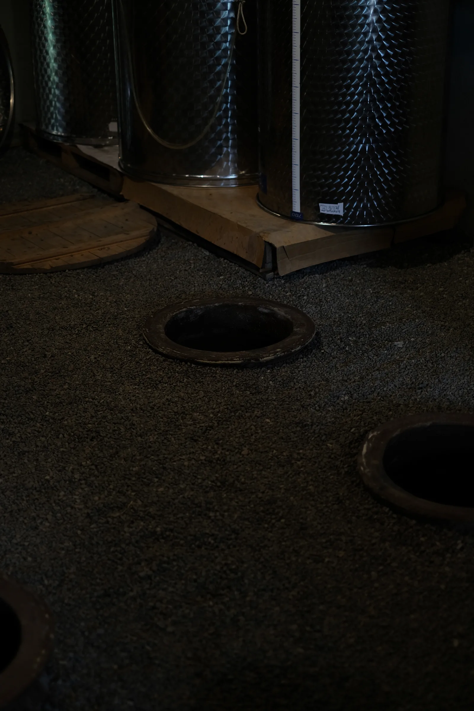 Open qvevri set into the gravel cellar floor, with stainless-steel tanks behind them.
