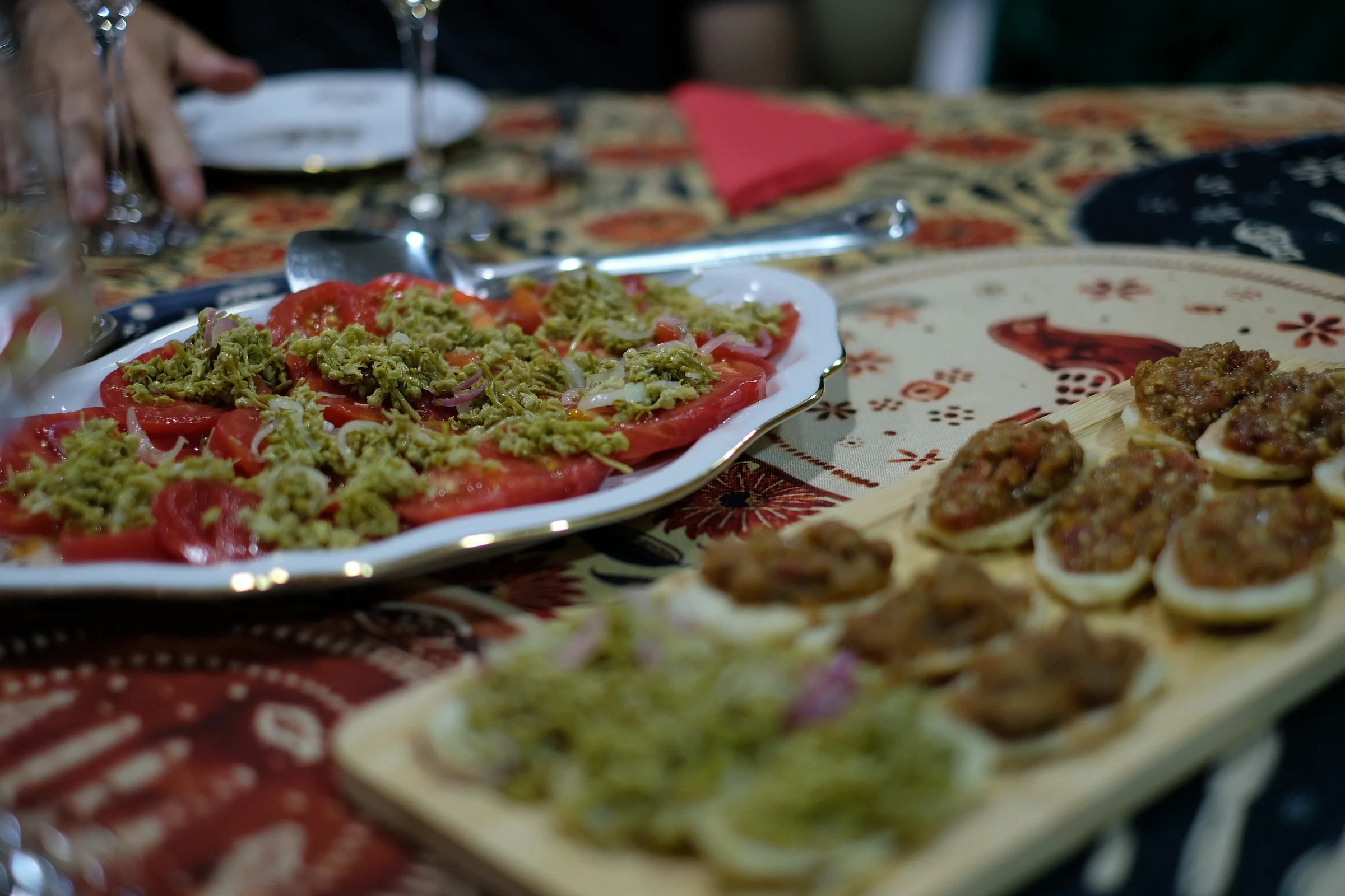 Tomatoes topped with jonjoli and red onion beside small toasts on a patterned tablecloth.